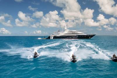 A large, luxurious yacht is seen in the background, surrounded by a vibrant turquoise ocean and fluffy white clouds in the sky. In the foreground, several people are being pulled behind the yacht on what appear to be motorized surfboards, creating a wake in the water.