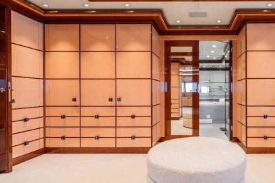 A spacious and well-designed walk-in closet with wooden cabinetry, recessed lighting, and a plush white ottoman in the foreground.