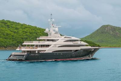 A large, luxurious yacht is anchored in a serene, tropical bay surrounded by lush, green hills and a clear, blue sky.
