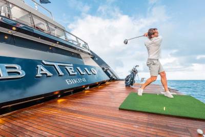 A person in casual attire is standing on a wooden deck of a large yacht, holding a golf club and preparing to take a swing, with the yacht's name "Batello" prominently displayed on the side.