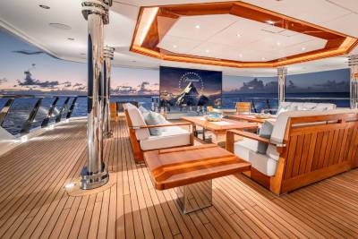 A luxurious outdoor deck area on a yacht, featuring comfortable seating, a wooden floor, and a scenic view of a city skyline in the background.