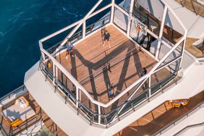 The image shows a wooden deck on the side of a ship, with people walking on it and the deep blue ocean visible in the background.