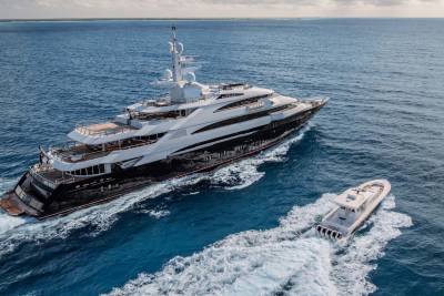 A large, luxurious yacht sailing across the open ocean, accompanied by a smaller speedboat in the foreground, with a vast expanse of blue water and sky in the background.