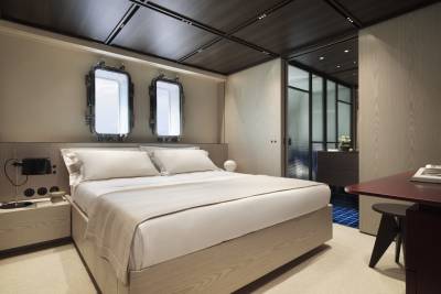 A modern bedroom features a light wood bed and minimalist decor, with porthole-style windows offering a glimpse of the outside.