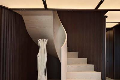 A sculptural staircase with light wood treads curves elegantly against dark, ribbed walls, illuminated by subtle ceiling lights.