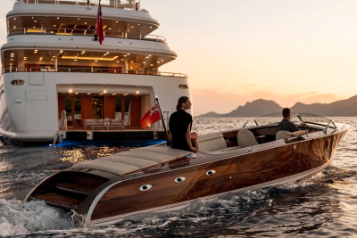 A sleek wooden speedboat glides through the water near a luxurious yacht at sunset.