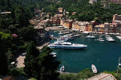 A picturesque Italian harbor is filled with colorful buildings, lush green hills, and luxurious yachts.
