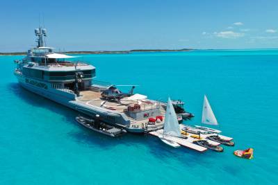A luxurious superyacht anchors in crystal-clear turquoise waters, surrounded by an array of watercraft and a helicopter.