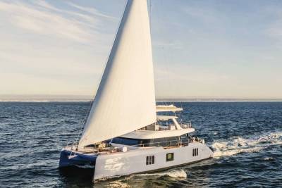A large white sailboat with a tall mast glides across the calm blue waters, with a scenic horizon visible in the background.