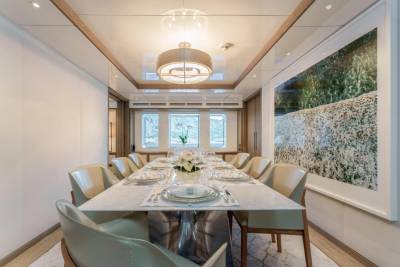 A long marble dining table is set for a formal meal aboard a luxurious yacht, adorned with a floral centerpiece and overlooking a scenic ocean view.