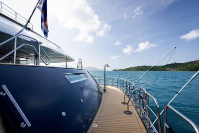 A large, sleek yacht with a wooden deck and railing is anchored in a serene, turquoise-colored body of water surrounded by lush, hilly terrain under a partly cloudy sky.