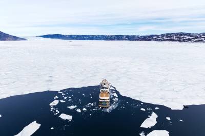 A small ship is seen floating amidst the frozen expanse of a vast, snow-covered landscape, with distant mountains visible in the background.