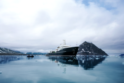 A large, dark-colored ship is reflected in the calm, icy waters of a fjord, with a snow-capped mountain peak rising in the background against a cloudy sky.