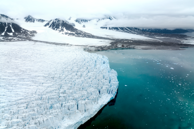 A massive glacier with a jagged, icy surface extends into a turquoise-colored body of water, surrounded by snow-capped mountains shrouded in mist.