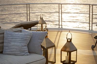 A cozy outdoor seating area on a boat or ship, with plush gray pillows and lanterns overlooking the serene water and sunset in the background.