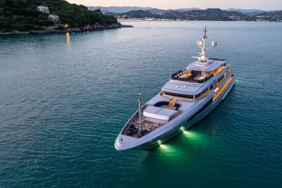 A large, luxurious yacht is anchored in a serene, turquoise bay surrounded by lush, verdant hills and buildings in the distance.