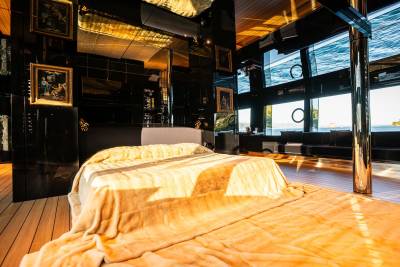 A cozy and luxurious bedroom with a large bed draped in a warm, golden blanket, surrounded by dark wooden furniture and framed artwork, with a panoramic window offering a breathtaking view of the outdoors.