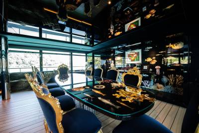 A cozy and eclectic dining room with a large wooden table surrounded by plush chairs, adorned with various decorative elements and framed artwork on the walls, and large windows offering a scenic view of the outdoors.