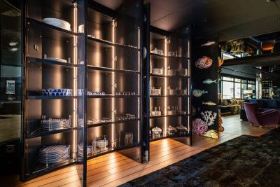 A dimly lit interior space with a large glass-enclosed shelving unit displaying various glassware and decorative items, set against a backdrop of a cozy lounge area with plush seating.