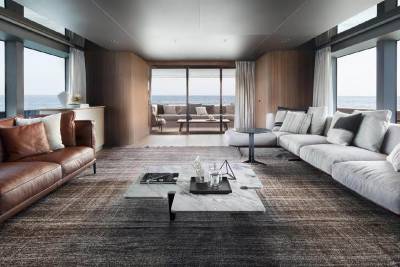 A modern and spacious living room with large windows overlooking a scenic view, featuring a leather sofa, plush armchairs, and a glass-topped coffee table on a patterned rug.