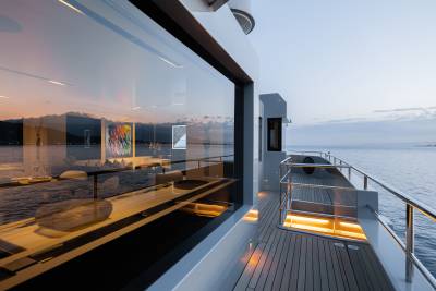 A modern, glass-enclosed balcony or deck overlooking a scenic body of water, with a wooden floor and various furnishings and decor elements visible.