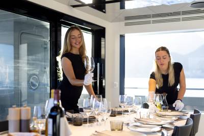 The image shows two women in black outfits standing at a table set for a formal dining event, with wine glasses, plates, and other tableware visible in the foreground.