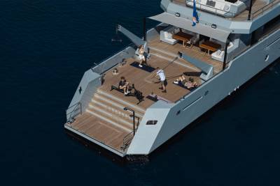 A large yacht with a wooden deck and people relaxing on the upper deck, surrounded by a dark blue body of water.