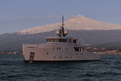 A large, gray yacht is floating on the calm waters of a bay, with a snow-capped mountain peak visible in the background.