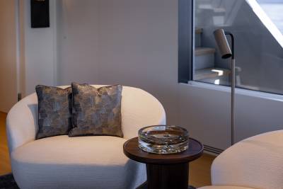 A cozy living room setting with a comfortable armchair, decorative pillows, and a glass ashtray on a wooden side table, set against a backdrop of a window and a wall.