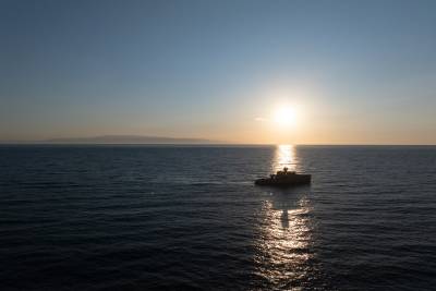 A small boat is silhouetted against the setting sun, which casts a warm glow over the calm, reflective waters of the vast ocean in the background.