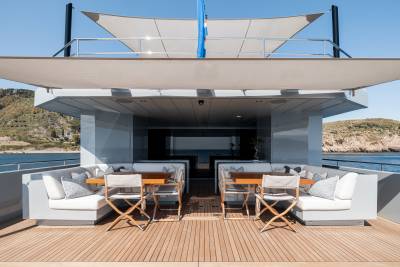 A modern, sleek yacht with a spacious outdoor deck featuring comfortable seating arrangements and a shaded canopy, set against a backdrop of a scenic coastal landscape.