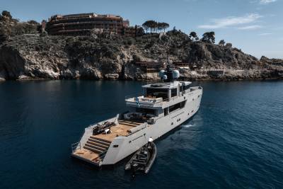 A large white yacht is anchored in a scenic cove surrounded by rugged cliffs and buildings perched on the hillside.