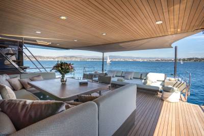 A luxurious yacht deck with comfortable seating and a dining table in the foreground overlooks a calm blue sea with sailboats and distant hills in the background.