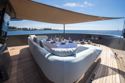 A luxurious outdoor dining and lounging area on a yacht's deck, with a table set for a meal and a large curved sofa in the foreground, overlooks a calm blue sea and a distant coastline under a bright sky.