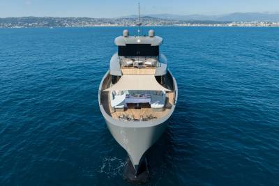 A sleek, modern yacht with an outdoor dining area in the foreground sails on calm blue water with a distant coastline in the background.