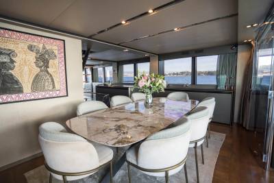 A modern dining area with a marble-topped table and plush chairs in the foreground, and a scenic view of the water and coastline through large windows in the background.