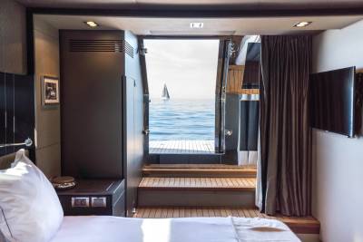 A luxurious yacht cabin with a bed in the foreground opens to a bright blue sea with a sailboat in the background.