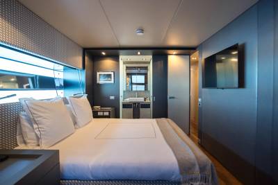 A luxurious yacht cabin features a neatly made bed in the foreground and a modern bathroom with a sink visible in the background.
