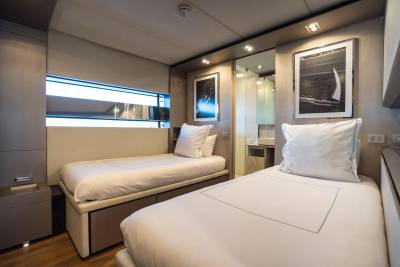 Two neatly made twin beds with white linens and pillows fill the foreground, while the background reveals a modern bathroom with a sink and a large mirror reflecting a sailboat image.