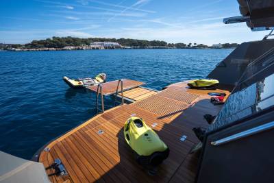 Two yellow underwater scooters rest on the wooden deck of a yacht, with a floating inflatable mat and a ladder extending into the calm blue water, and a tree-lined coast with buildings in the distance.