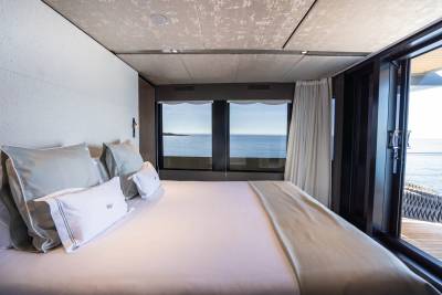 A neatly made bed with plush pillows occupies the foreground, while large windows in the background reveal a serene ocean view.