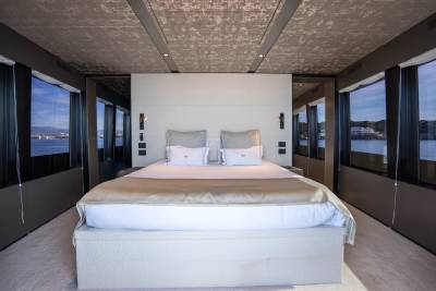 A luxurious yacht bedroom with a large bed in the foreground and large windows offering views of the water and distant coastline in the background.