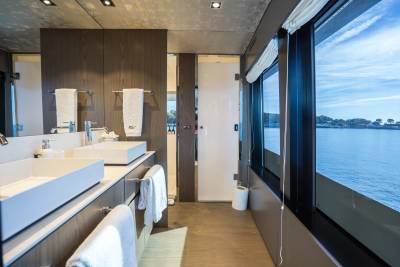 A modern bathroom with dual sinks in the foreground and a scenic view of the water and coastline through large windows in the background.