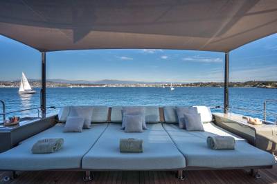 A spacious sun deck with plush cushions and rolled towels faces a tranquil blue sea dotted with sailboats and a distant, sun-drenched coastline.