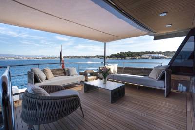 A modern yacht deck with comfortable seating in the foreground overlooks a calm blue sea and a distant coastline under a clear sky.