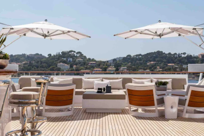 Two large white umbrellas shade a luxurious outdoor seating area on a yacht's deck, with a lush, green hillside dotted with houses in the background.