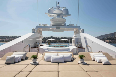A luxurious yacht with plush seating in the foreground is anchored in a calm bay with a lush, green hillside in the background.