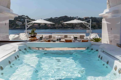 A luxurious jacuzzi with clear blue water sits in the foreground, while a sun-drenched deck with lounge furniture and umbrellas overlooks a picturesque coastline in the background.