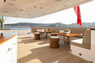 A luxurious yacht deck with comfortable seating and tables in the foreground overlooks a sun-drenched coastline with a red flag flying in the background.