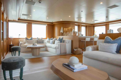 Plush white sofas and a modern coffee table fill the foreground of a luxurious yacht lounge, while a sleek bar area with stools and large windows form the background.